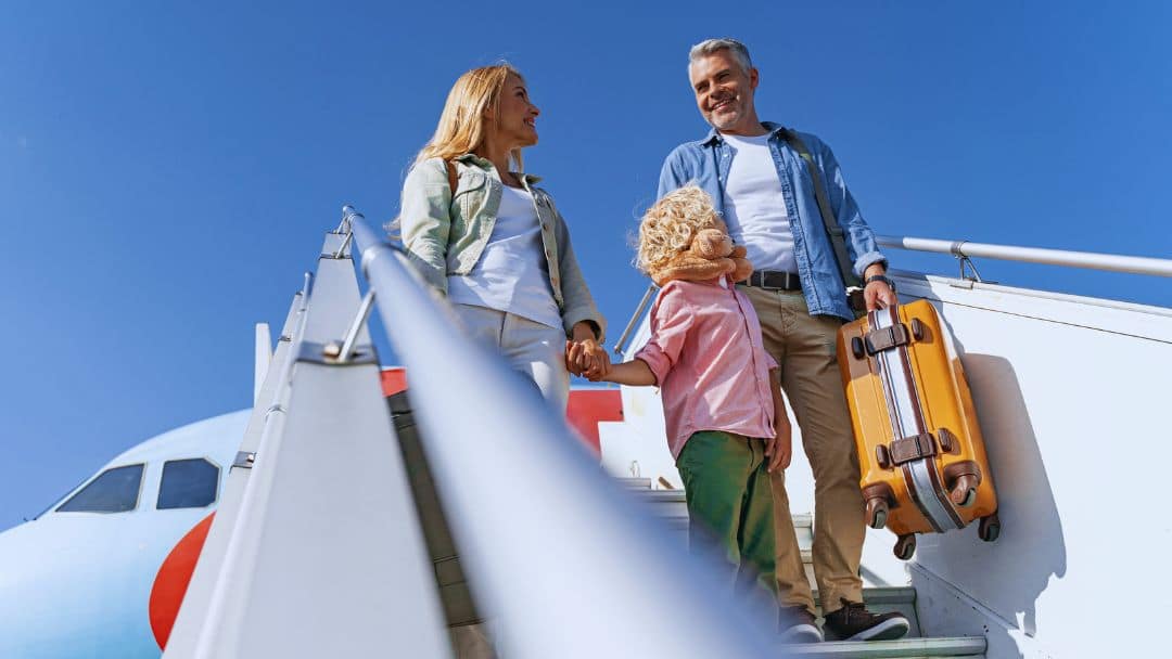 Happy family – mom, dad, and young child – boarding an airplane with luggage under blue sky.