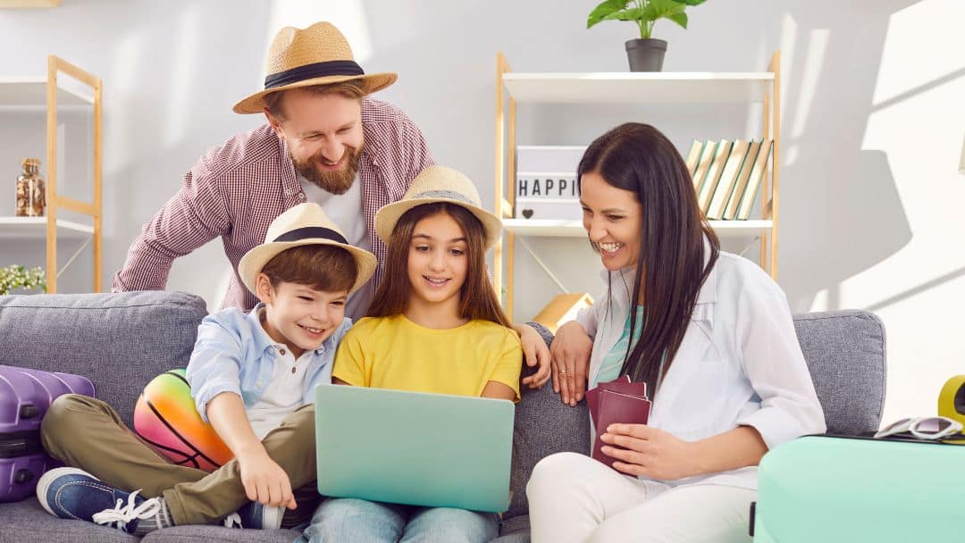 A family of four, including a mother, father, daughter, and son, is sitting on a couch in a bright living room, looking at a laptop. They are wearing straw hats and are surrounded by luggage, suggesting they are planning or booking a family vacation.