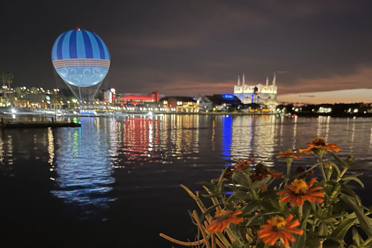 Night image of a Disney hot air balloon over a lake with colorful lights reflecting, featuring a castle and flowers in the foreground, showcasing a magical evening scene.
